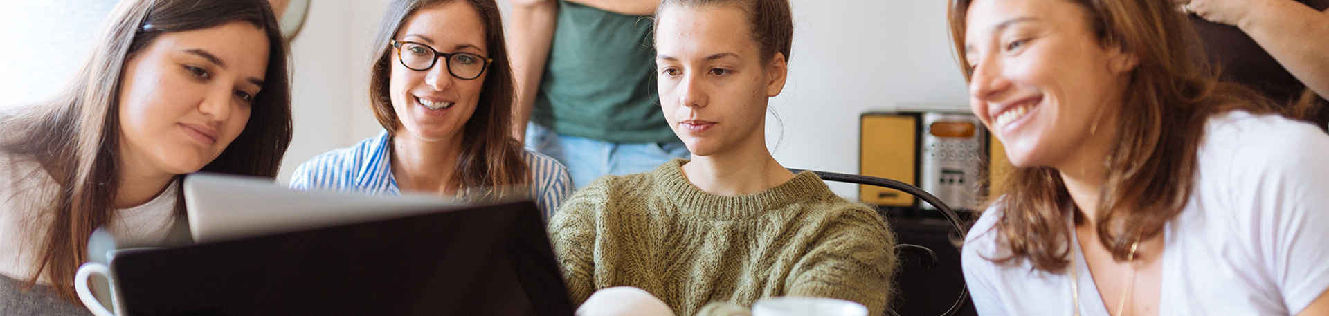 Eine Gruppe von Personen sitzt eng beisammen und schaut auf einen Laptop-Bildschirm. Im Hintergrund sind weitere Menschen und Möbelstücke, darunter ein Lautsprecher, sichtbar.